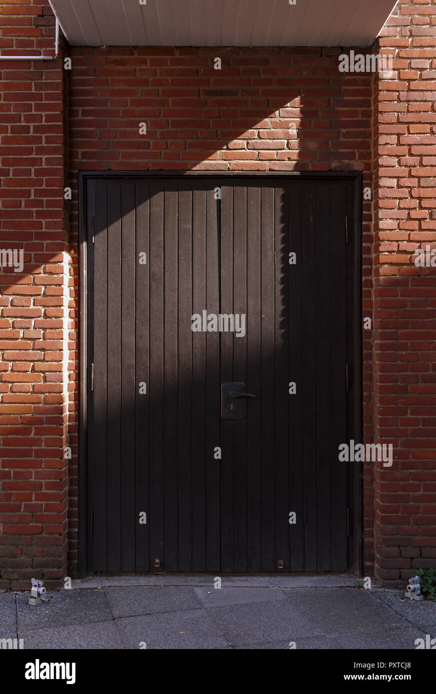 Sunlit red brick wall with dark wooden double leaved door and diagonal hard shadow Stock Photo
