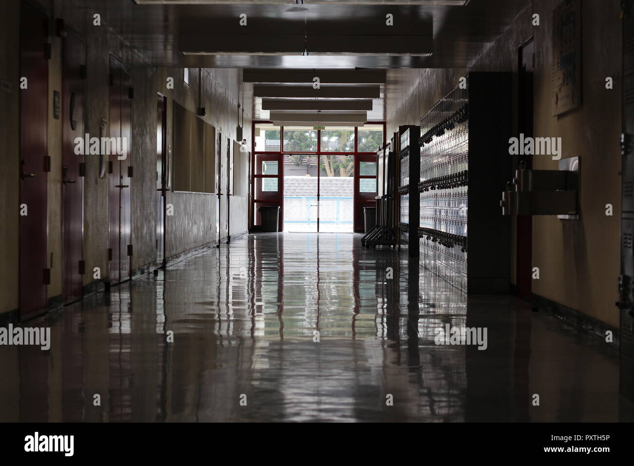 A dark and empty school hall and shiny floors during the day with lockers and class doors on each side and open red doors in the background Stock Photo