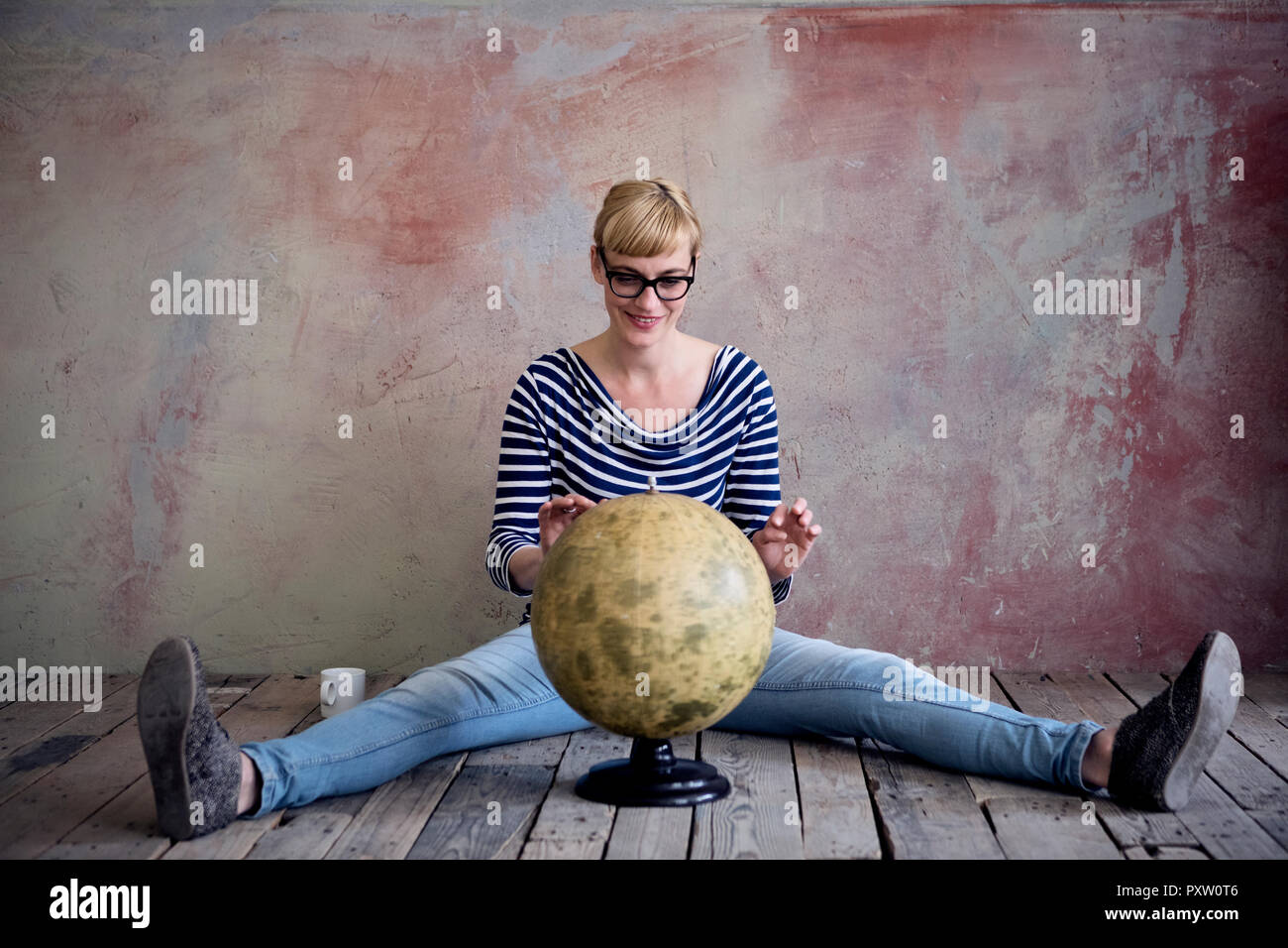 Smiling woman sitting on wooden floor in an unrenovated room looking at globe Stock Photo