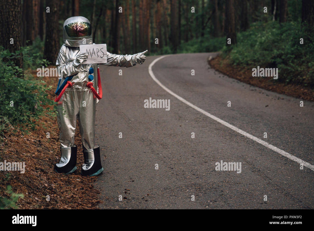 Spaceman hitchhiking to Mars, standing on road in forest Stock Photo