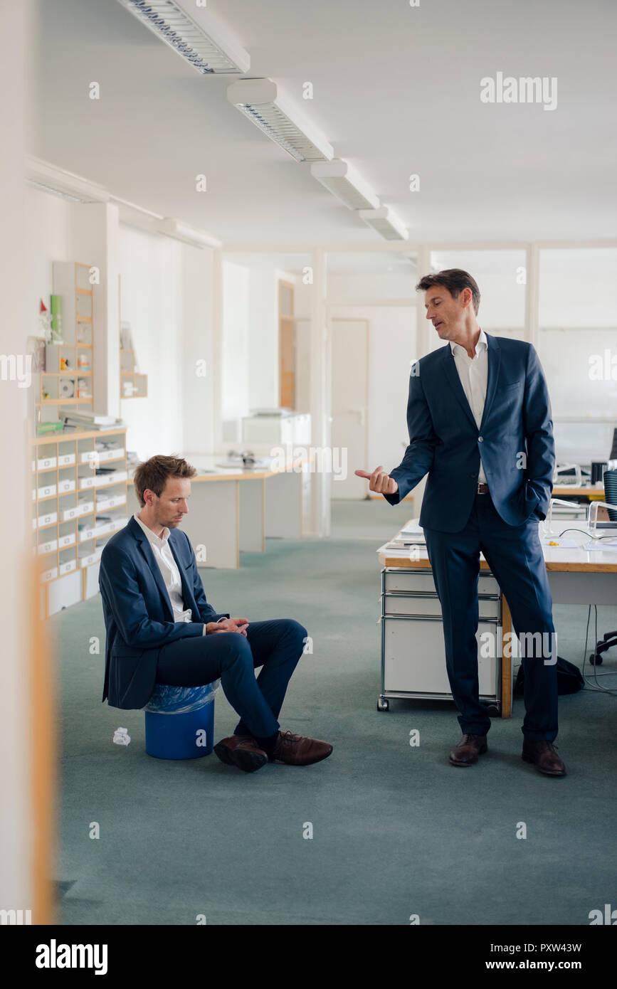 Manager making colleague, sitting on wastepaper basket, redundant Stock Photo