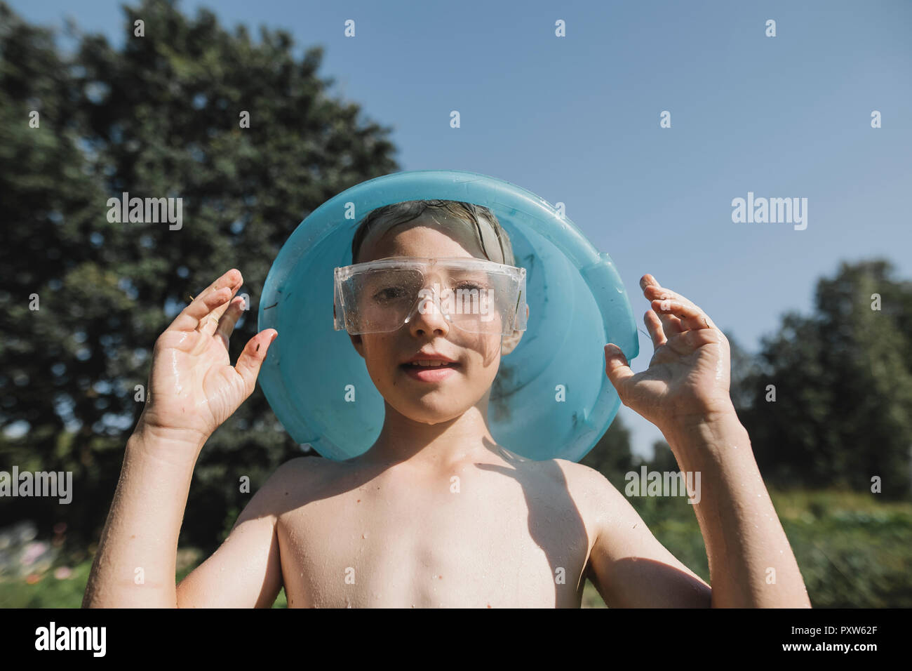 Wet boy wearing safety goggles holding bowl above his head in garden Stock Photo