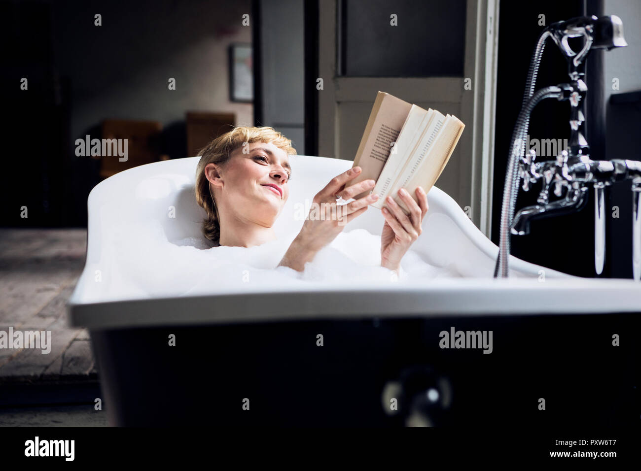 Portrait of relaxed woman taking bubble bath in a loft reading a book Stock Photo