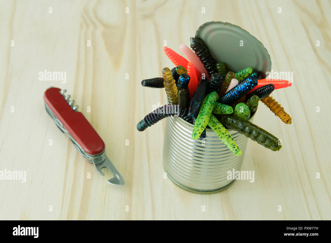 Colourful weird artificial plastic worms crawling out of tin can opened with pocket knife on wooden background Stock Photo