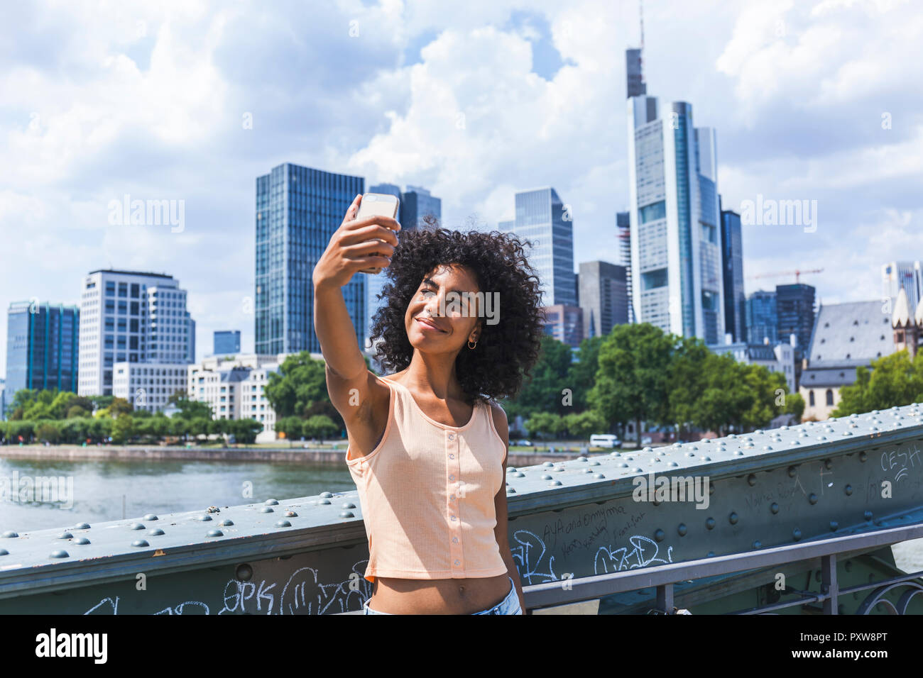 Germany, Frankfurt, portrait of content young woman with curly hair taking selfie in front of skyline Stock Photo