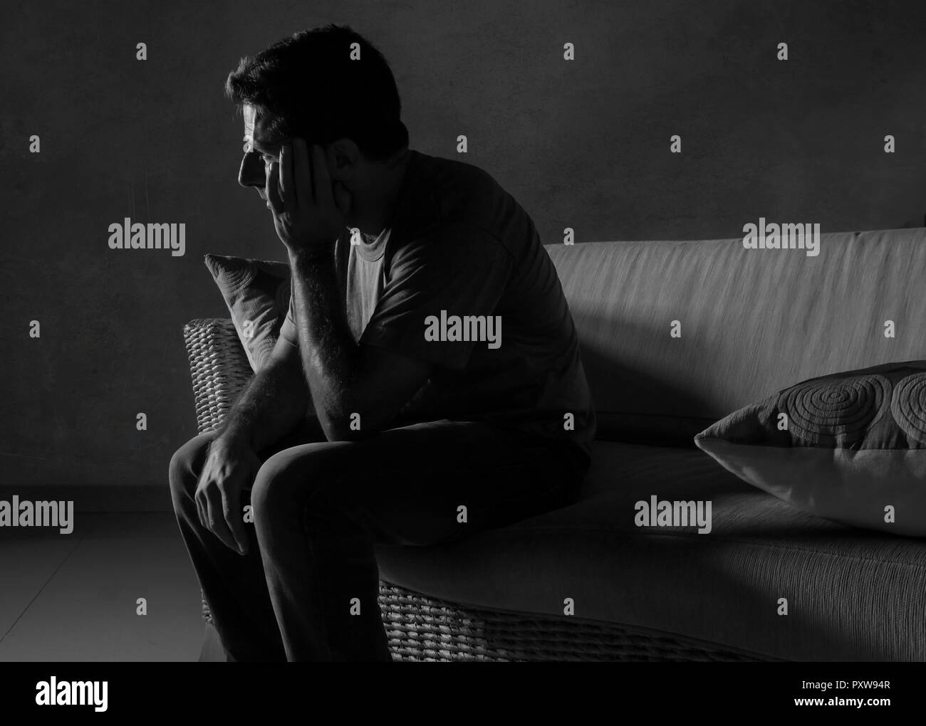 lifestyle dramatic light portrait of young sad and depressed man sitting at shady home couch in pain and depression feeling lost looking away thoughtf Stock Photo