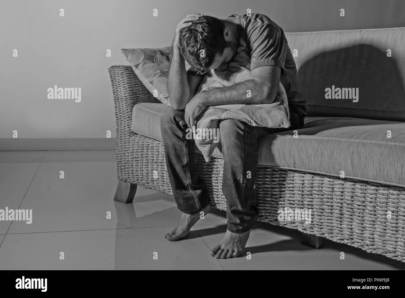 lifestyle dramatic light portrait of young sad and depressed man sitting at shady home couch in pain and depression feeling stressed and desperate cry Stock Photo