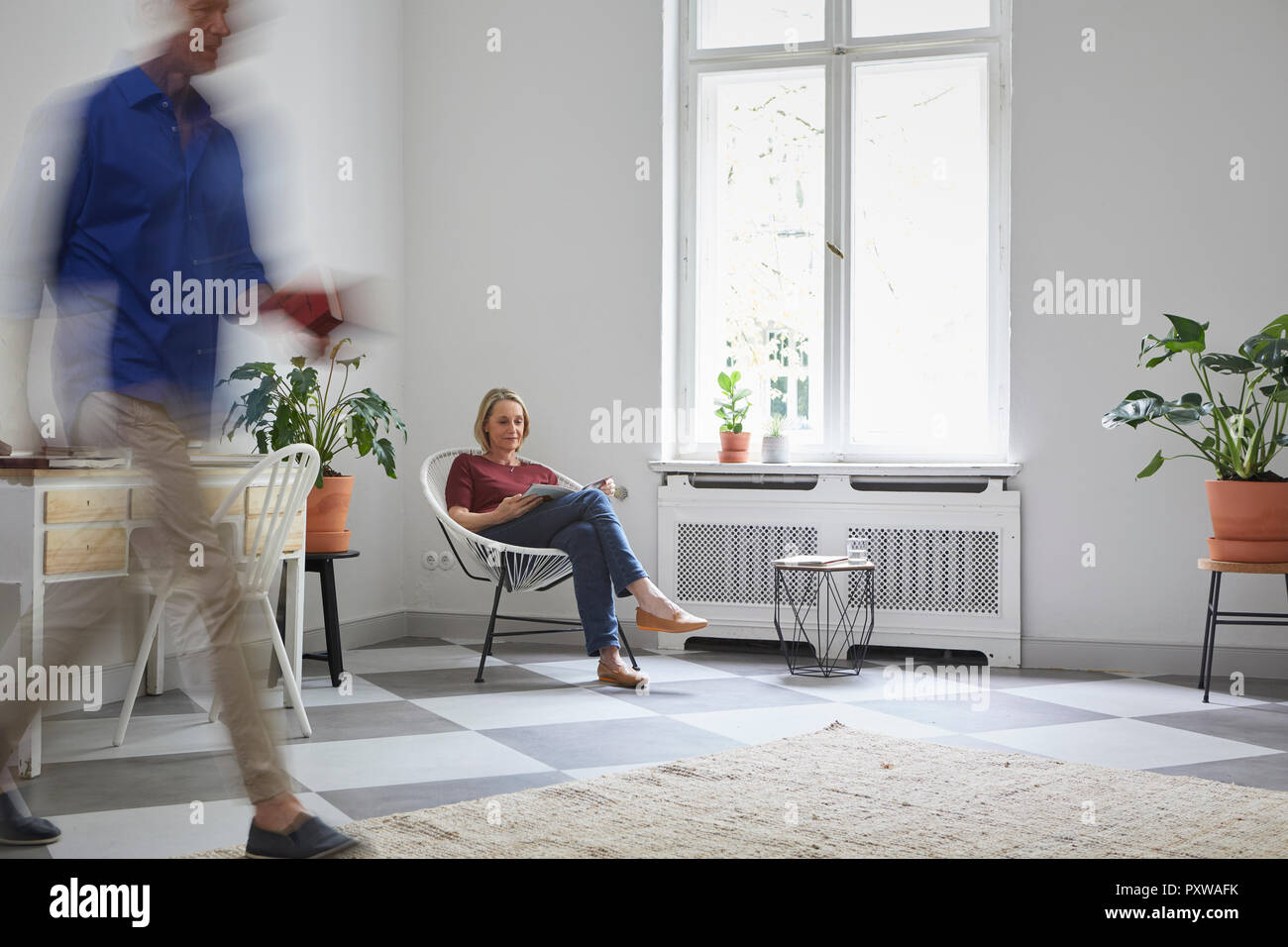 Mature woman reading magazine at home with man passing by Stock Photo