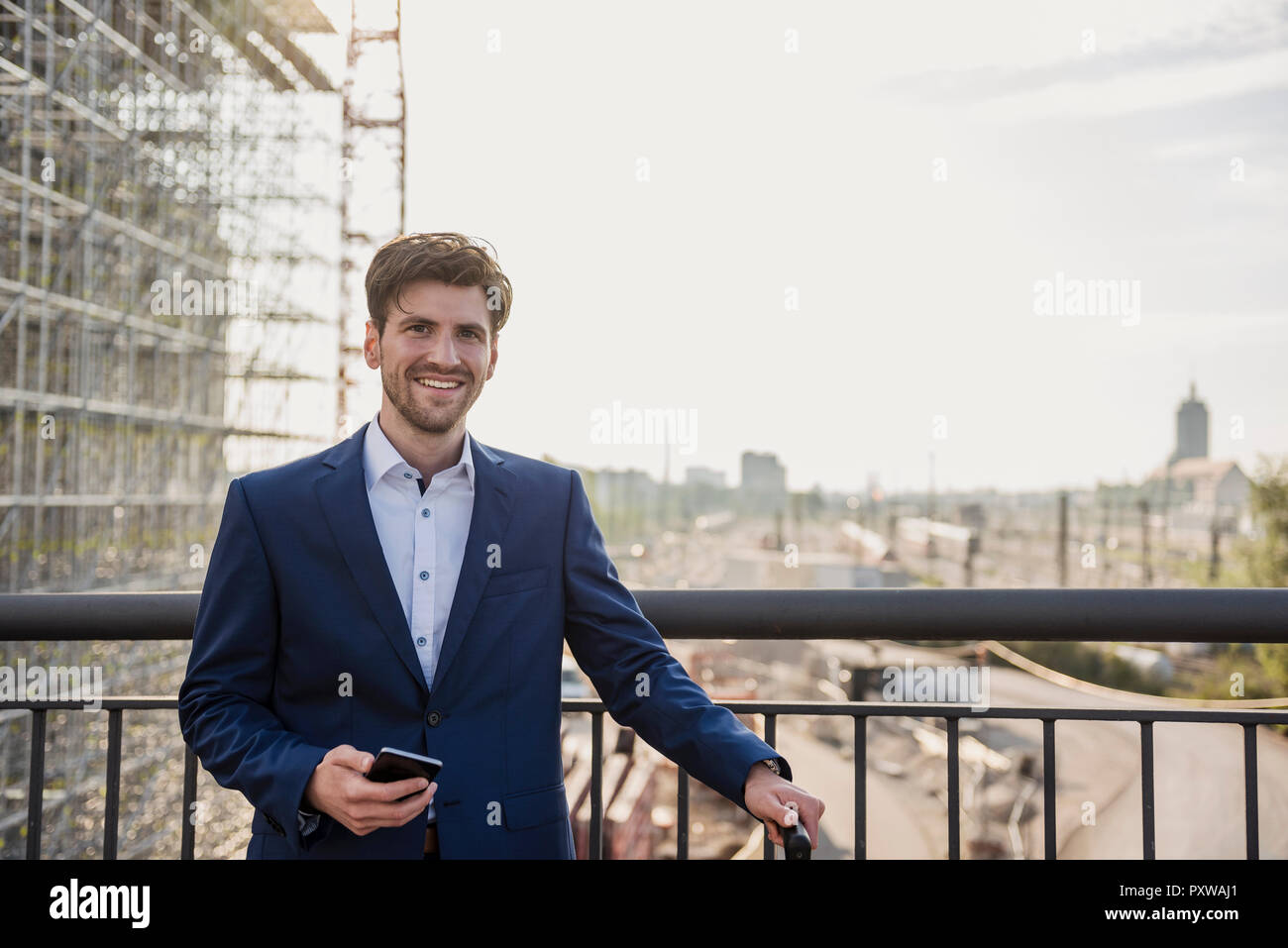 Portrait of smiling businessman standing on bridge in the city holding cell phone Stock Photo