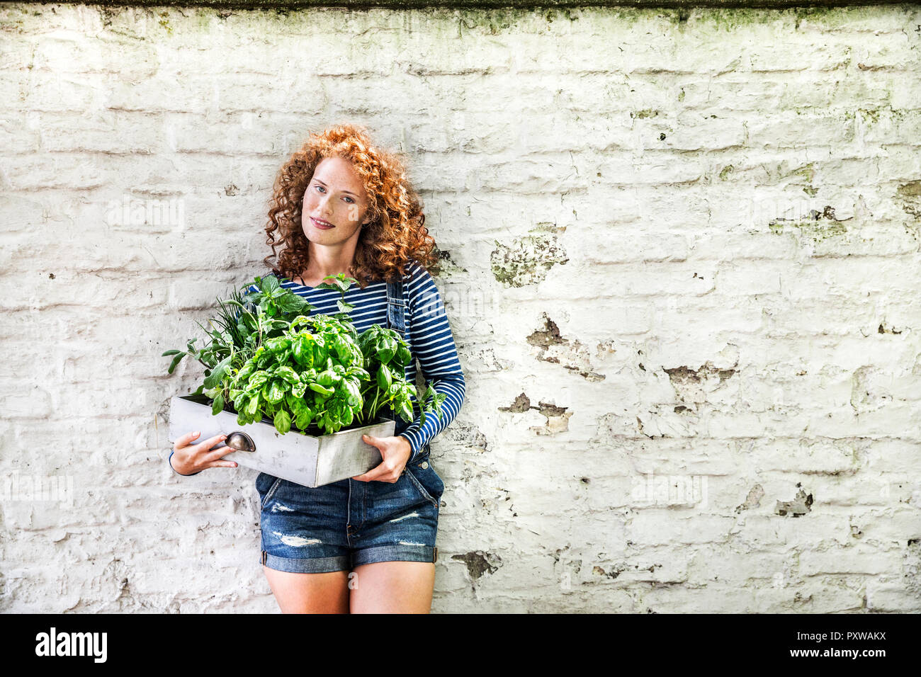 Portrait of young woman with fresh herbs in a box leaning against white brick wall Stock Photo