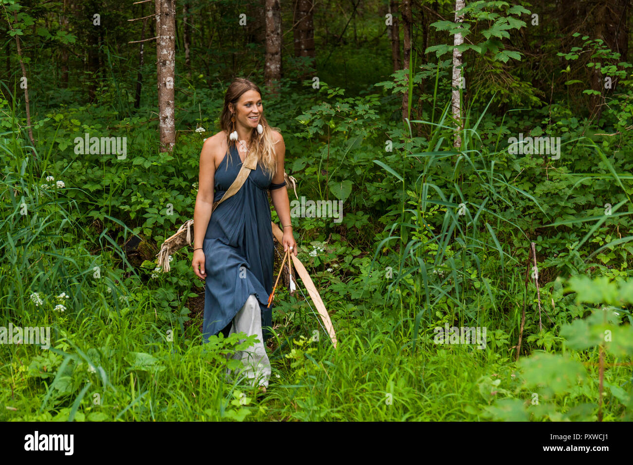 Smiling woman walking in a forest with bow and arrow Stock Photo
