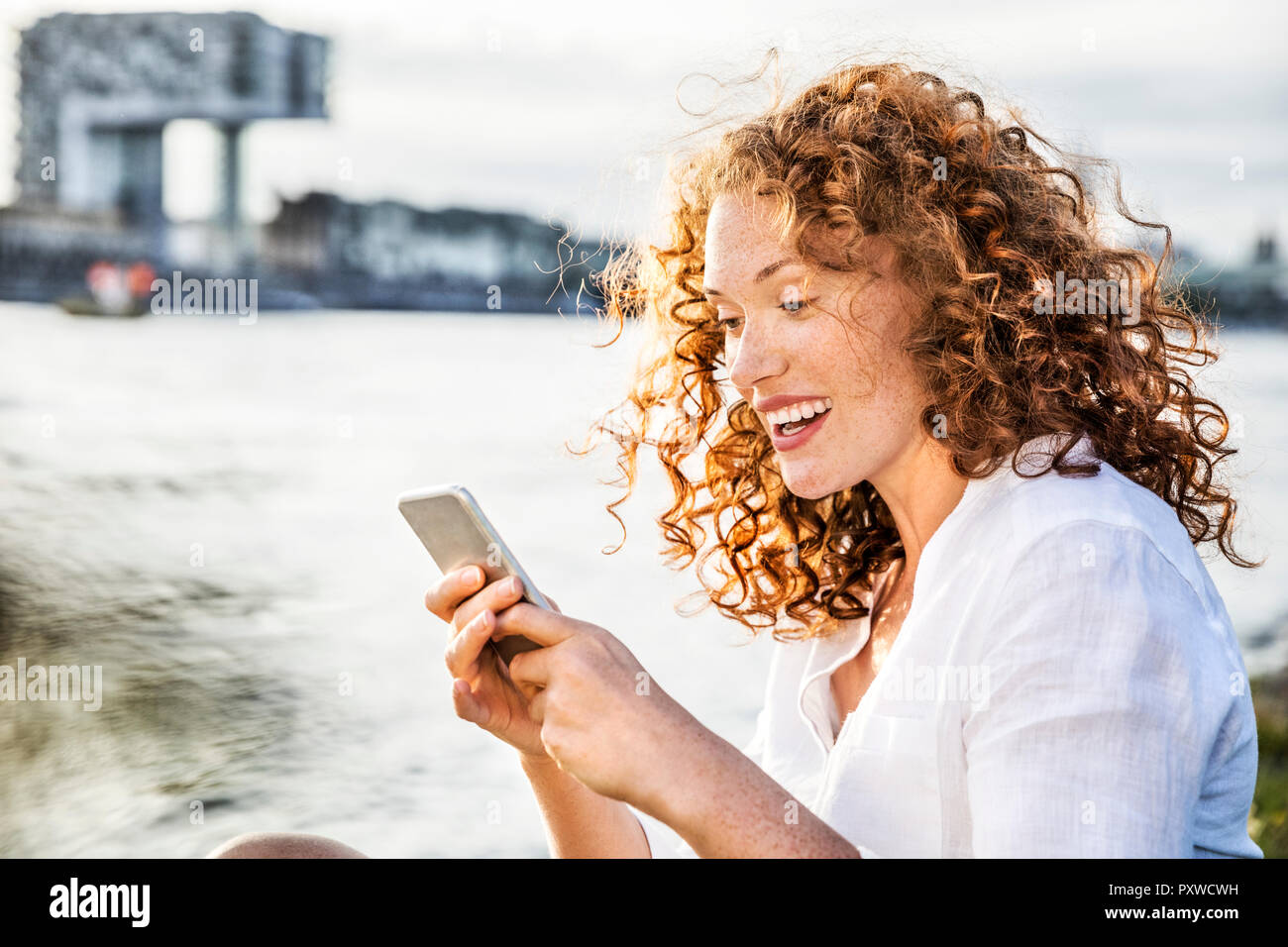 Germany, Cologne, portrait of amazed young woman sitting at riverside looking at cell phone Stock Photo