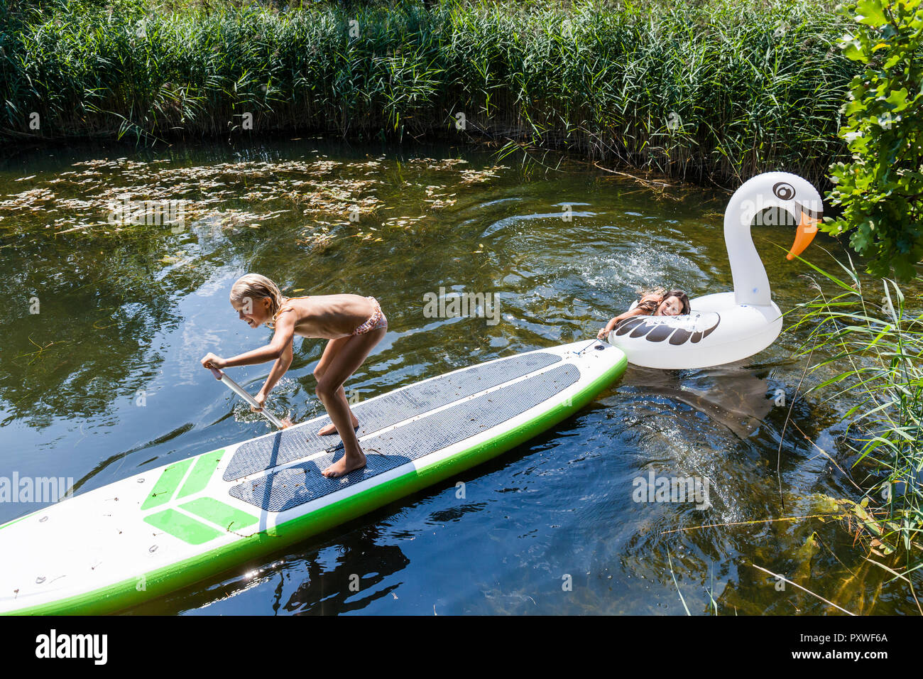 Two girls in a pond with inflatable pool toy in swan shape and SUP board Stock Photo