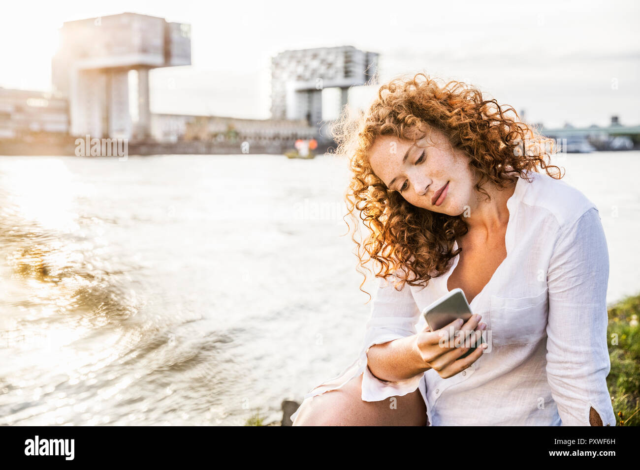 Germany, Cologne, portrait of young woman sitting at riverside looking at cell phone Stock Photo