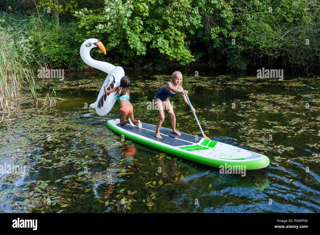 Two girls in a pond with inflatable pool toy in swan shape and SUP board Stock Photo