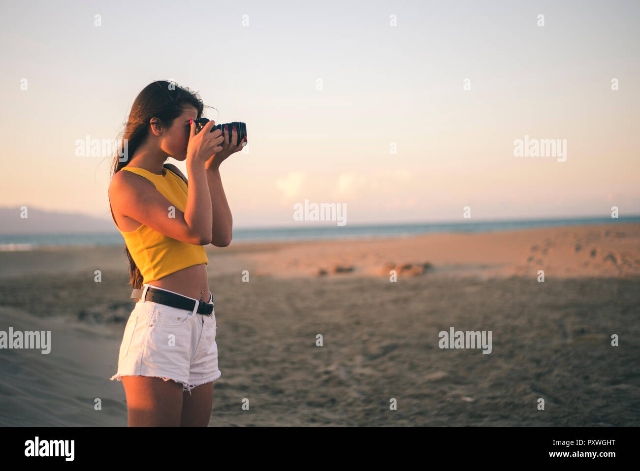 Teenage girl taking photos with camera on the beach at sunset Stock Photo