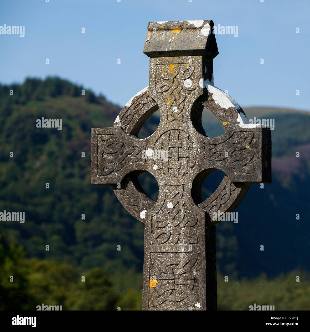 A carved, stone celtic cross in Ireland, with green hills and a blue sky behind. Moss is growing on the stonework which is old. Stock Photo