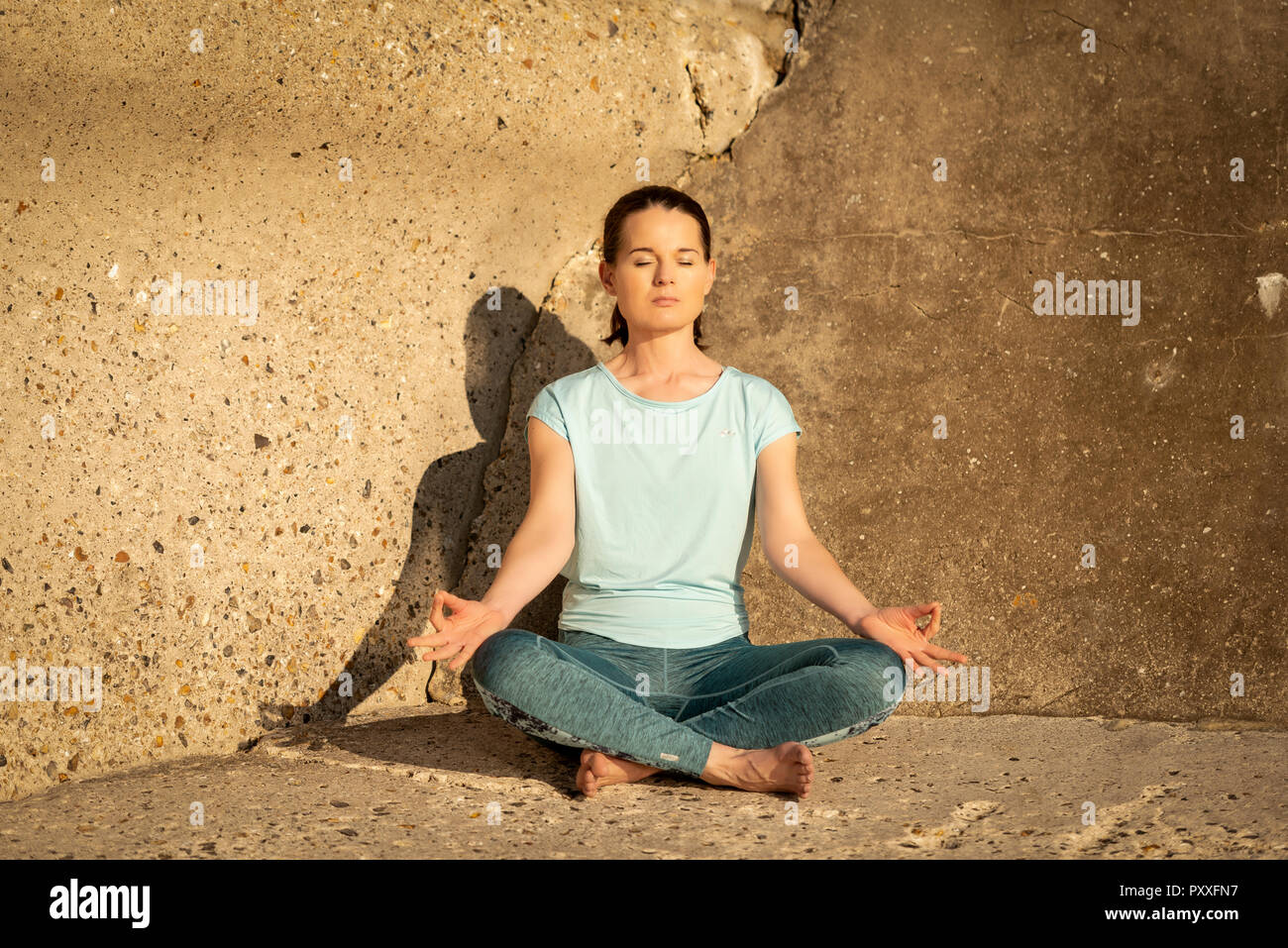 woman practicing yoga and meditating in the sun by a concrete wall Stock Photo