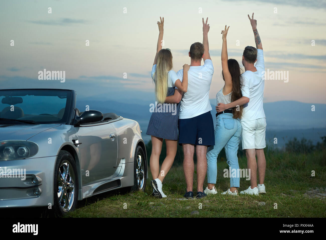 Back view of two couples standing, admiring amazing view and landscapes, holding hands up and showing peace. Man putting arms round girls' waists. Friends posing near silver cabriolet. Stock Photo