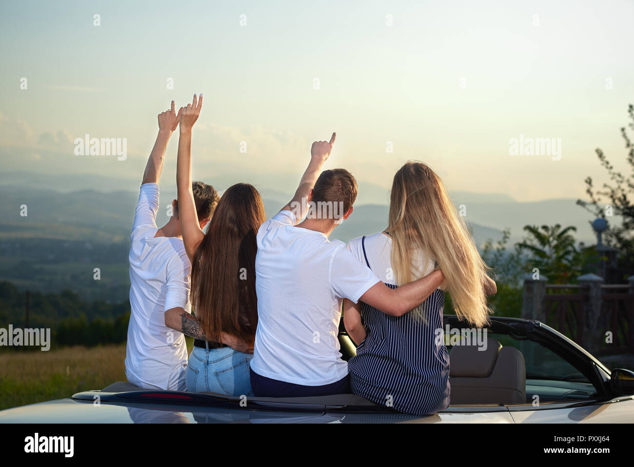 Two couples sitting on cabriolet and admiring landscapes and amazing view. Men putting arms round girls' waists and holding hands up. Friends enjoying summertime and journey by car. Stock Photo
