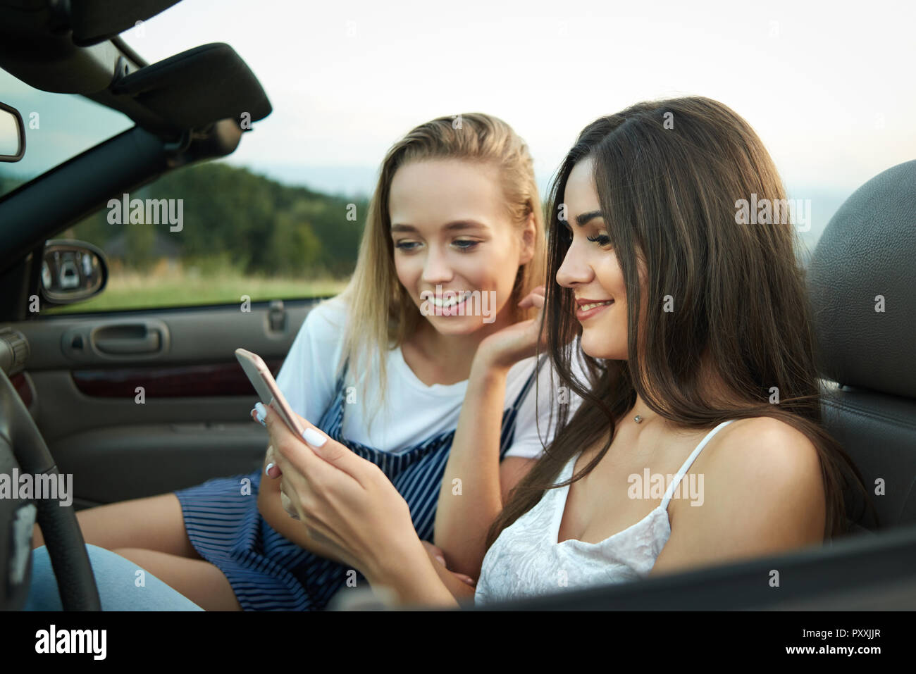 Beautiful brunette and blonde sitting on front saet of automobile. Two female friends posing in cabriolet and holding phone and looking at it. Girls spending time together on weekends. Stock Photo