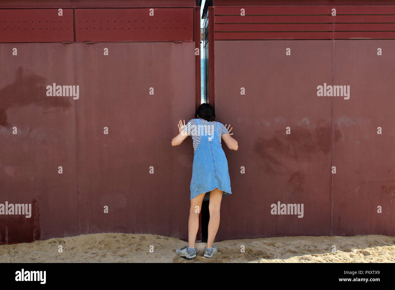 Girl at the beach peeping the hidden sea trough a slit in steel wall Stock Photo