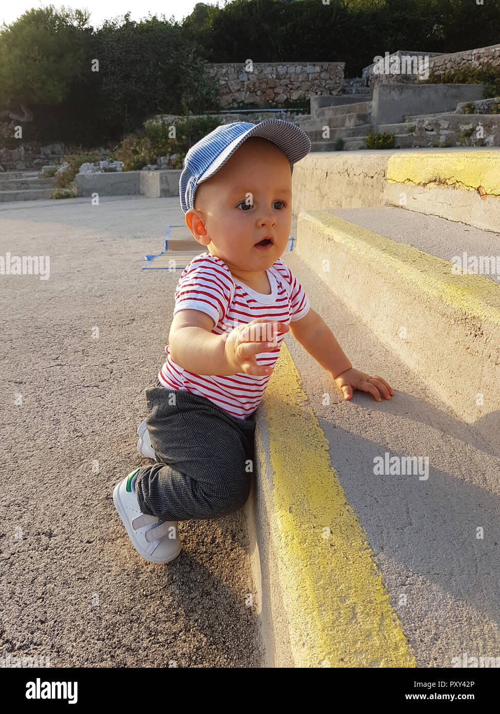 Cute Baby Boy 1 Year Wearing A Cap Backwards And Striped Clothes, Close Up Portrait, Baby Standing On The Stairs Stock Photo