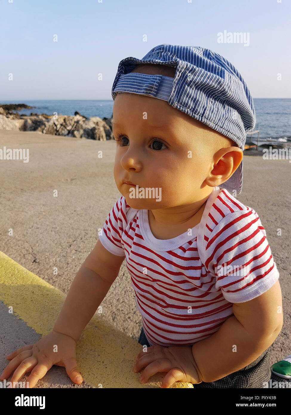 Cute Baby Boy 1 Year Wearing A Cap Backwards And Striped Clothes, Close Up Portrait. Mediterranean Sea In The Background Stock Photo