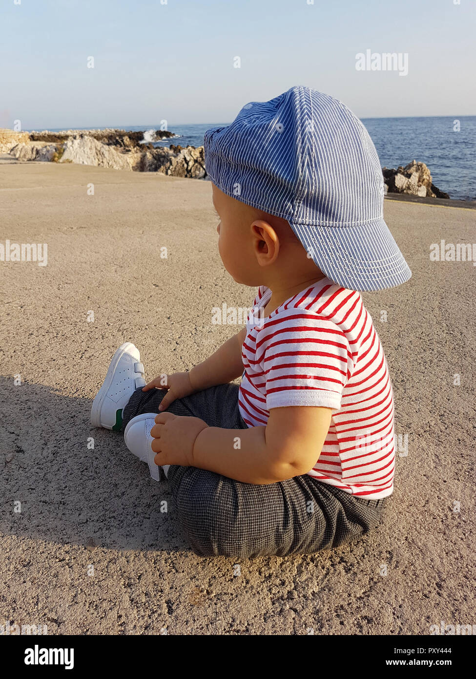 Cute Baby Boy One Year Wearing A Cap Backwards And Striped Clothes, Close Up Portrait. Mediterranean Sea In The Background Stock Photo