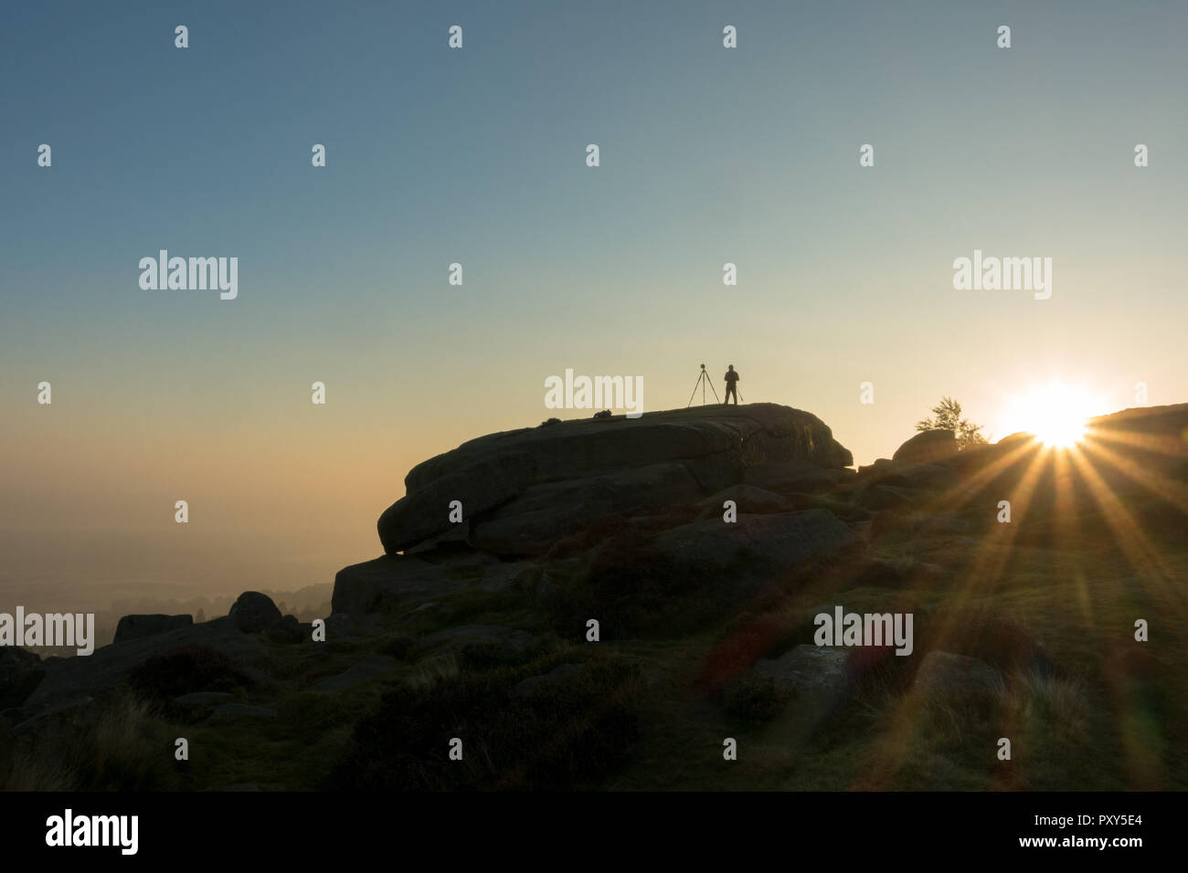 Silhouette of a photographer with a tripod set up taking landscape photos of the sunrise on Ilkley Moor's Cow and Calf Rocks, West Yorkshire, UK Stock Photo