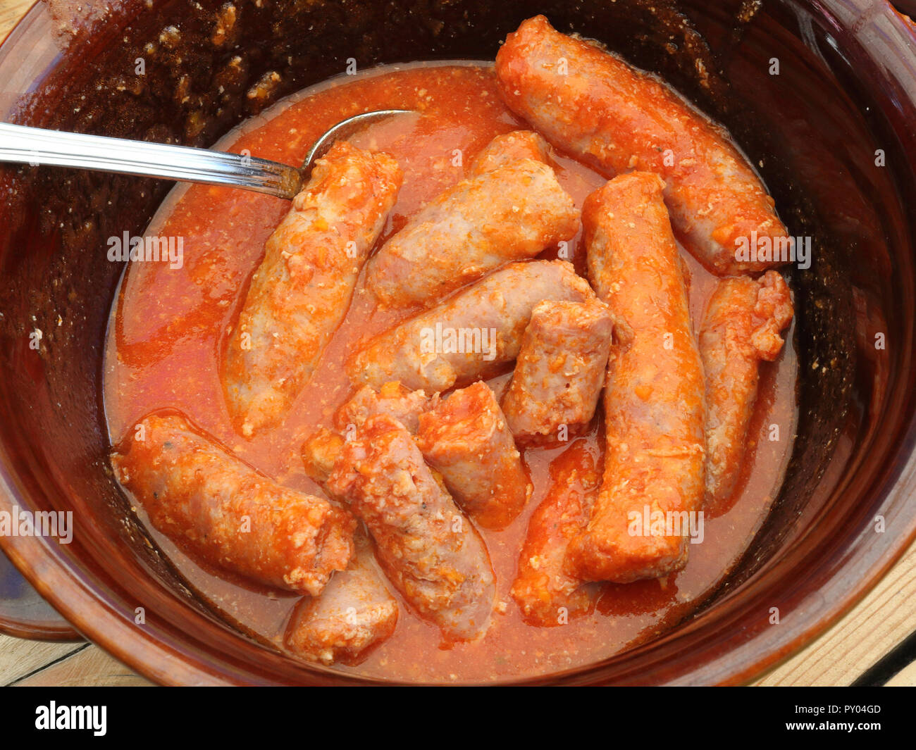 A brown bowl on a wooden table with a spoon containing a homemade sausages stew in spices and tomato sauce to eat with polenta, a typical Italian dish Stock Photo