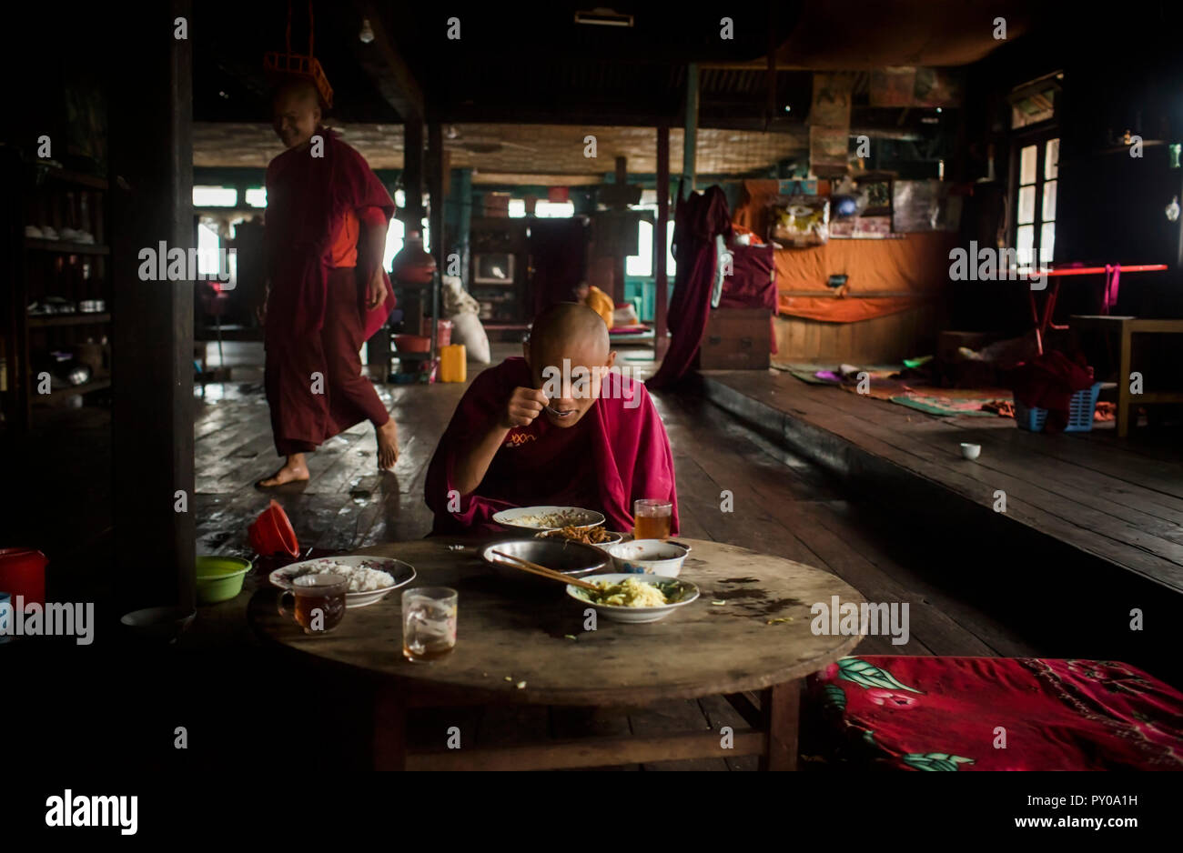 Young monk eating at table in rural monastery with another monk walking behind in background, Myanmar, Shan, Myanmar Stock Photo