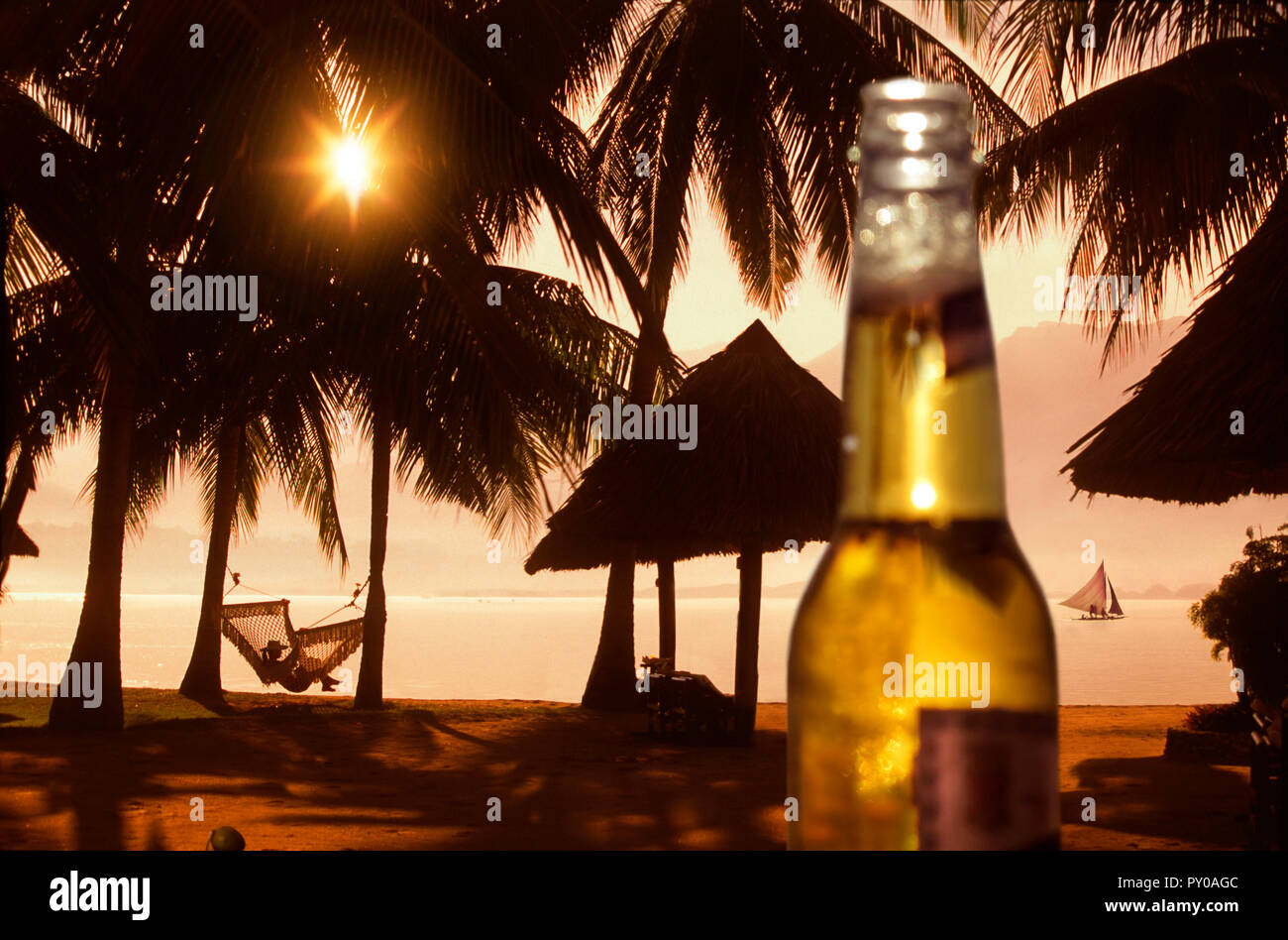 Beer bottle against beach with palm trees and woman in hammock at sunset, Badian, Cebu, Philippines Stock Photo