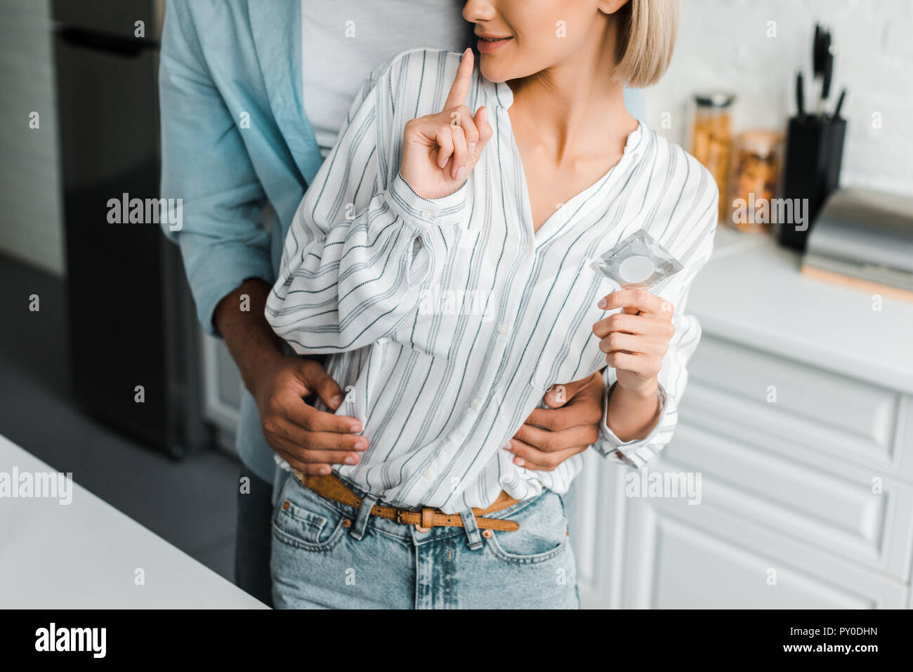cropped image of girlfriend showing silence gesture and holding condom in kitchen Stock Photo