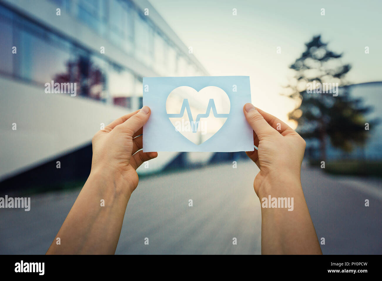 Close up of woman hands holding a paper sheet with the heart beating symbol inside, over hospital building background. Healthcare and medicine concept Stock Photo