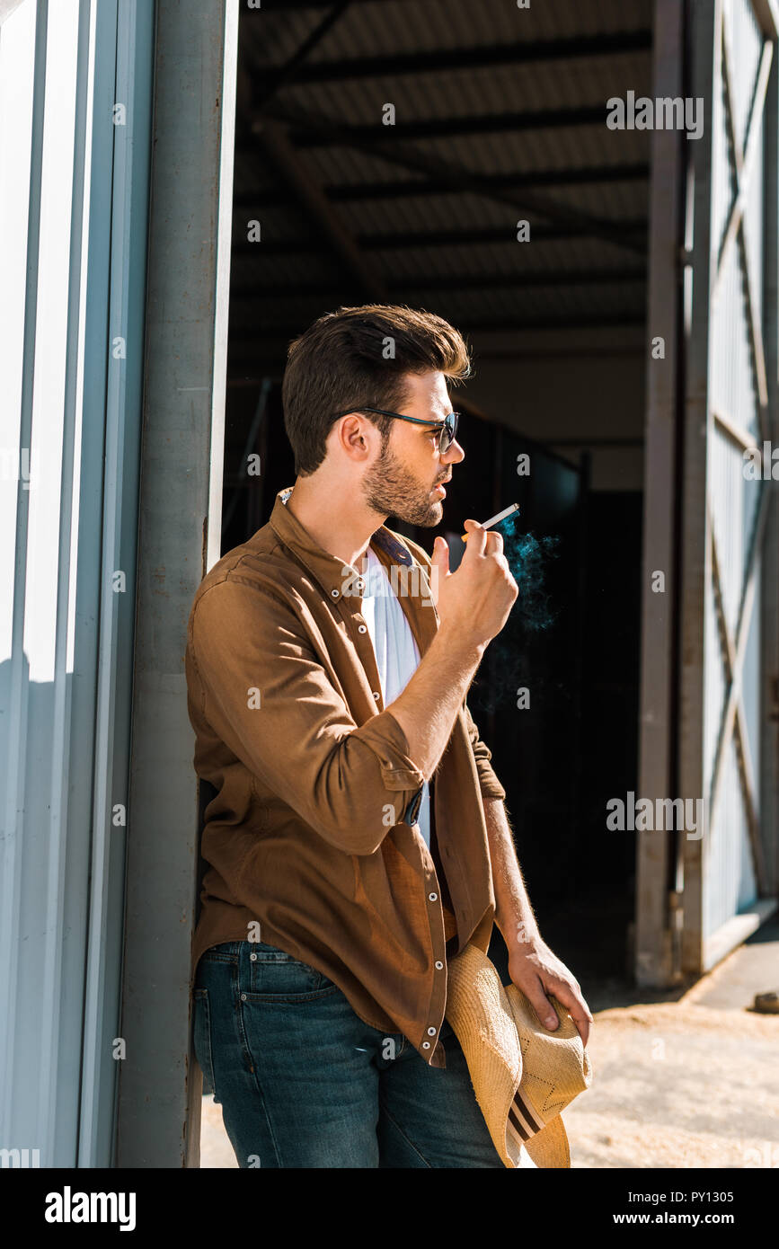 side view of handsome man in sunglasses smoking cigarette and leaning on wall at ranch Stock Photo