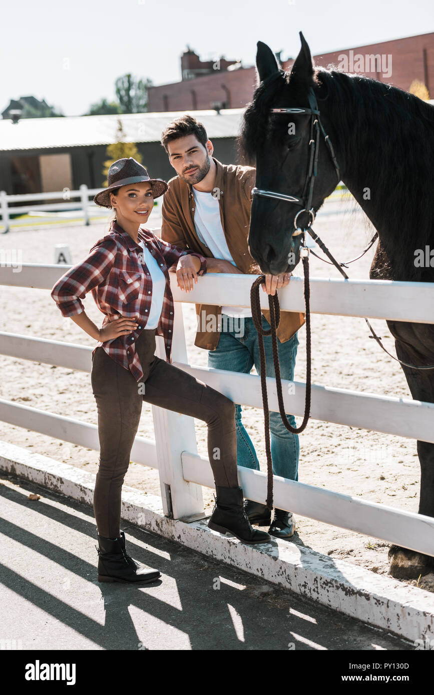 female and male equestrians standing near fence with horse at ranch and