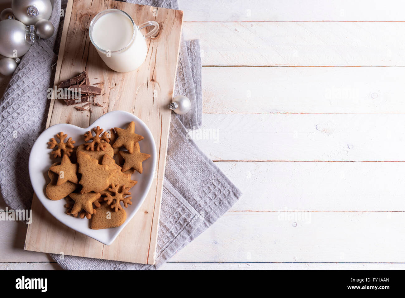 Santa Claus snack with gingerbread cookies on a heart-shaped plate, milk and chocolate, a kitchen towel and Christmas balls on a white table. Stock Photo