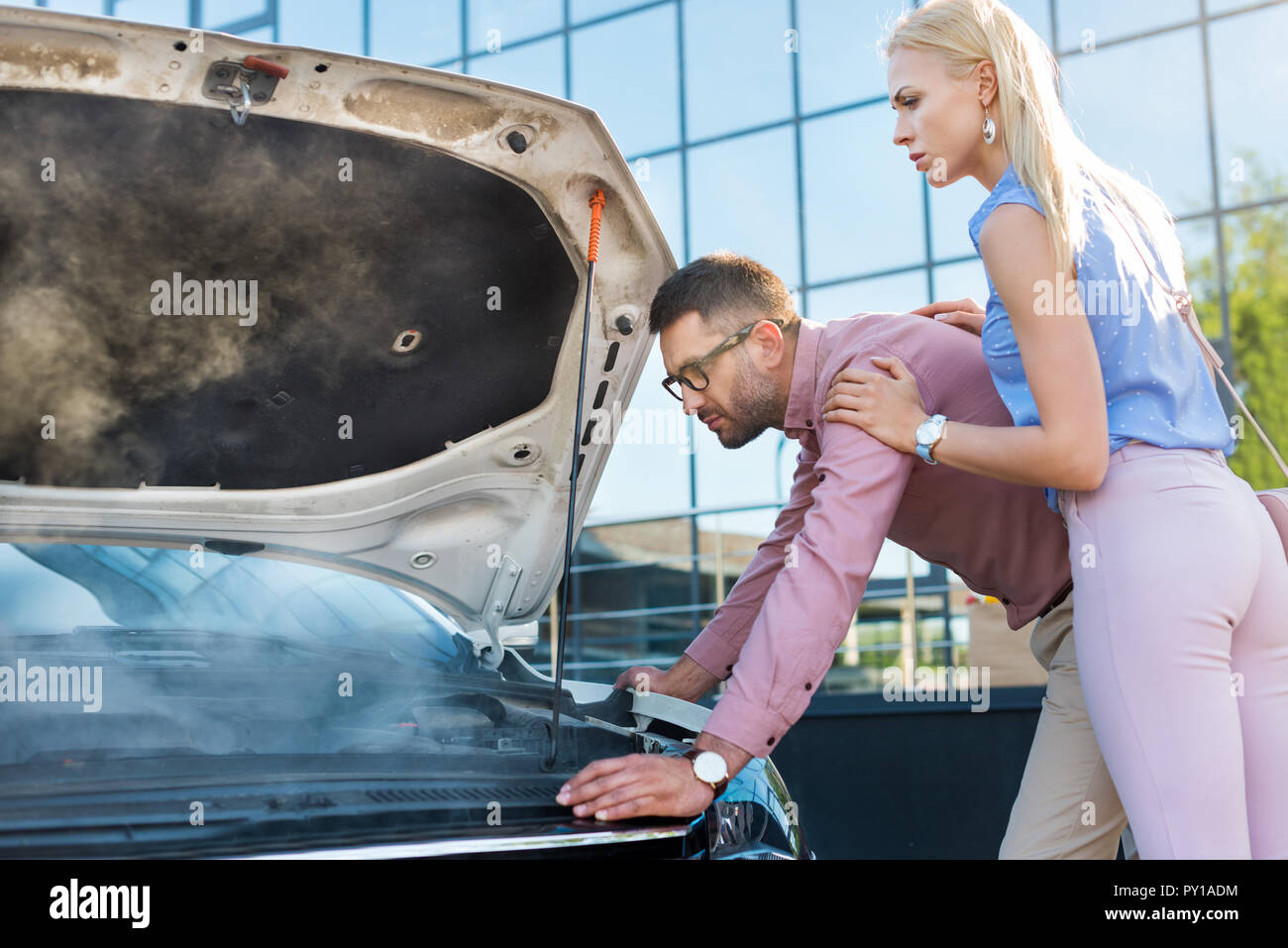 side view of couple looking under car hood of broken car on street