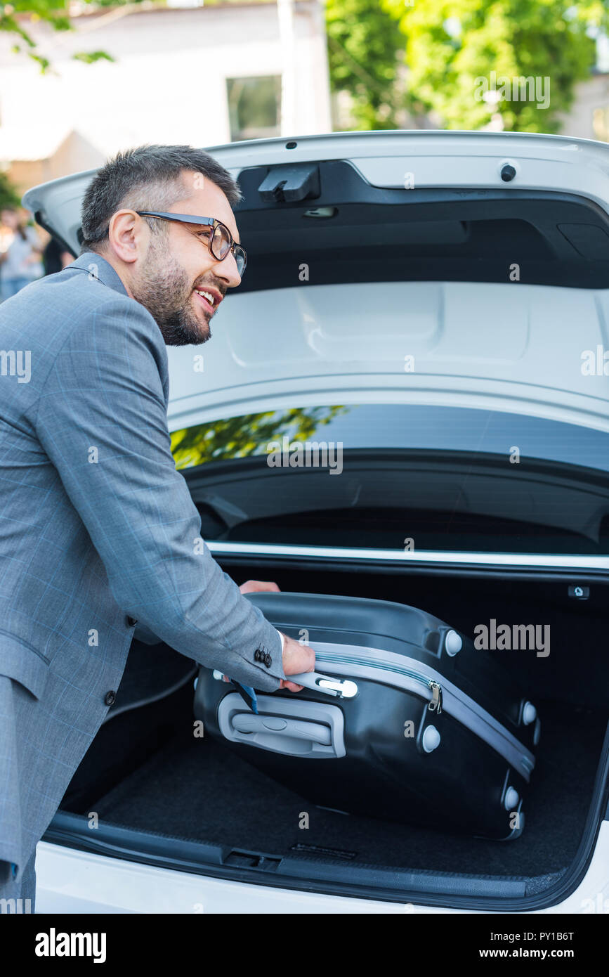 side view of smiling businessman putting luggage into car on parking ...