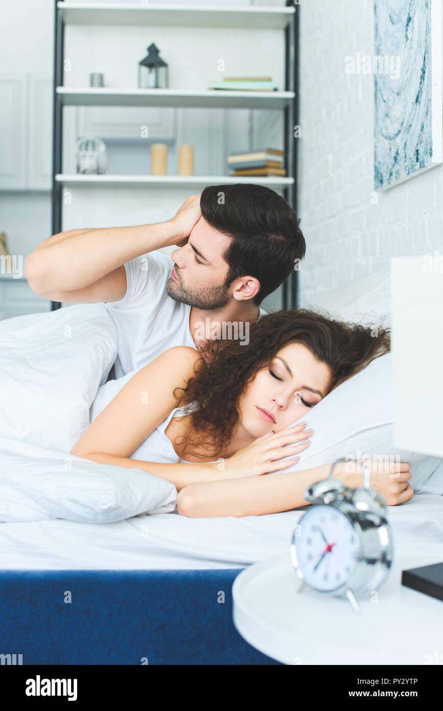 young man waking up and rubbing eye while girlfriend sleeping in bed Stock Photo
