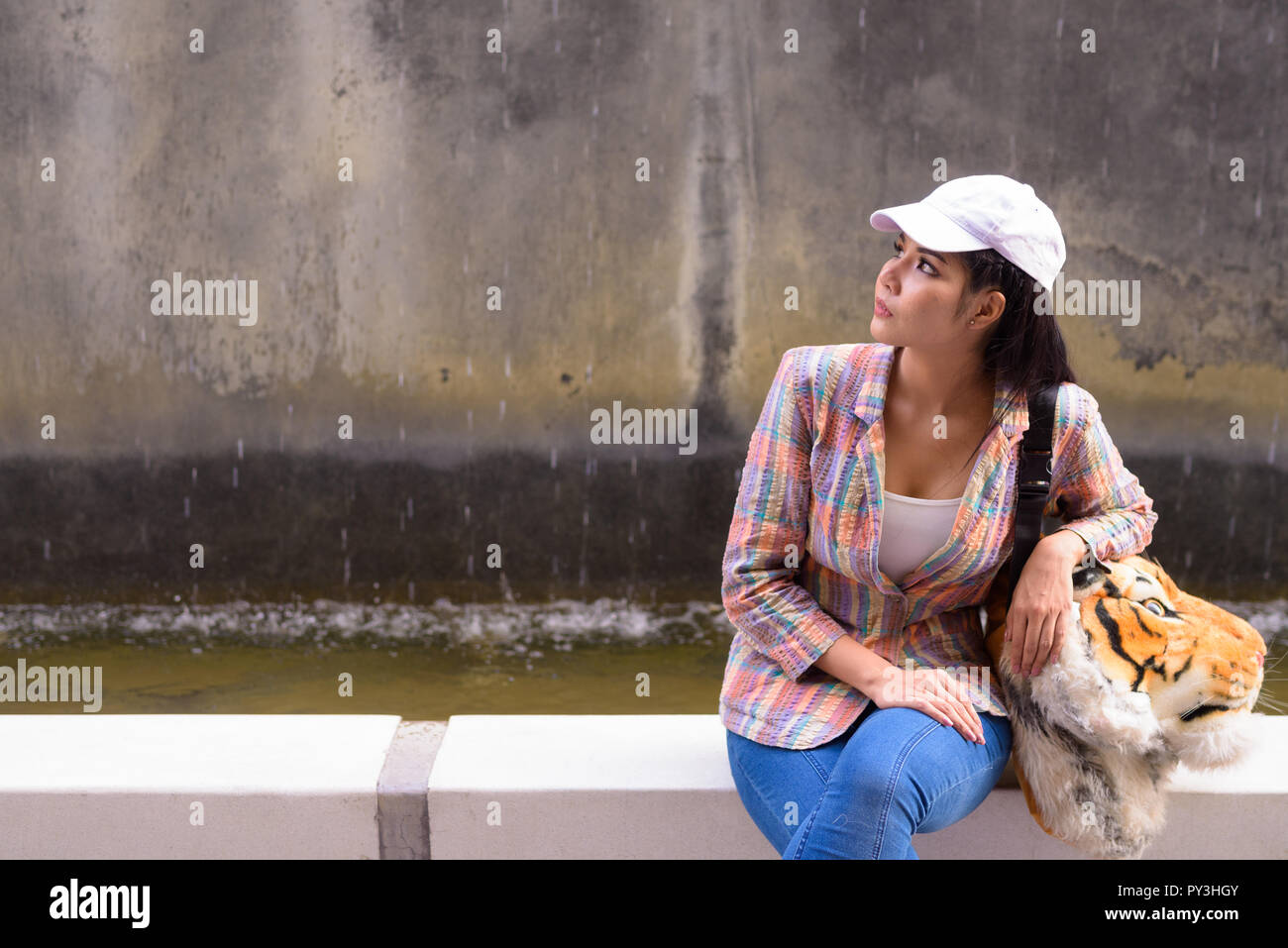 Portrait of young beautiful Asian woman sitting and thinking outdoors Stock Photo