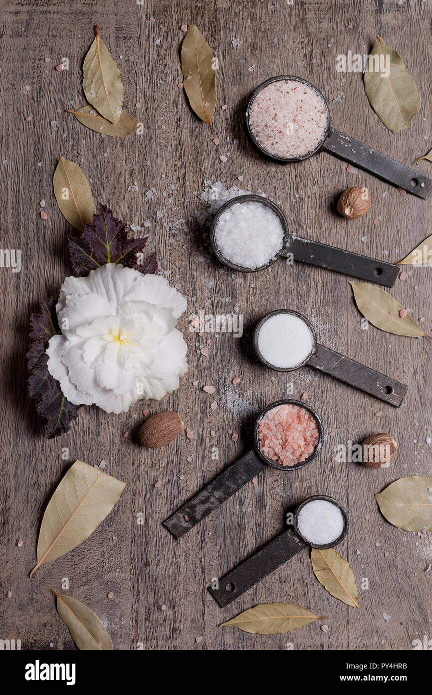 Still life of various types of salt on a wooden background with bay leaves, whole nutmeg and white begonia flower. Stock Photo