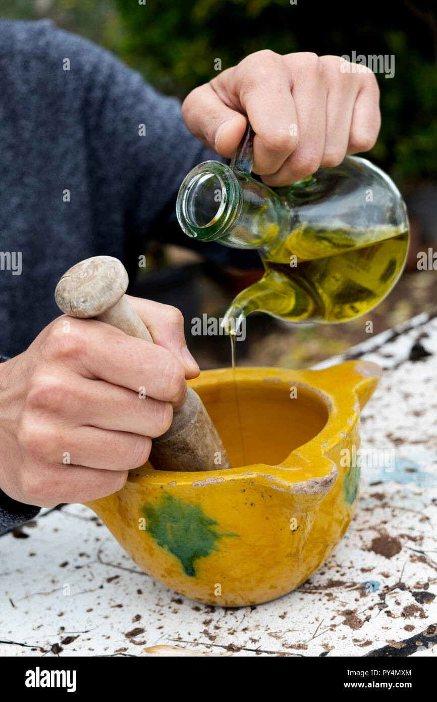 closeup of a young caucasian man outdoors making some aioli using a wooden pestle in a yellow ceramic mortar, by adding olive oil from a glass cruet Stock Photo