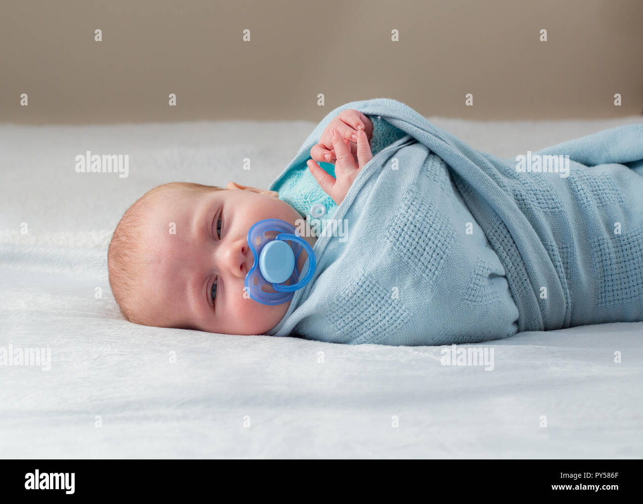 Baby laying down wrapped in a blue blanket facing the camera with sleepy eyes and pacifier in his mouth. Stock Photo