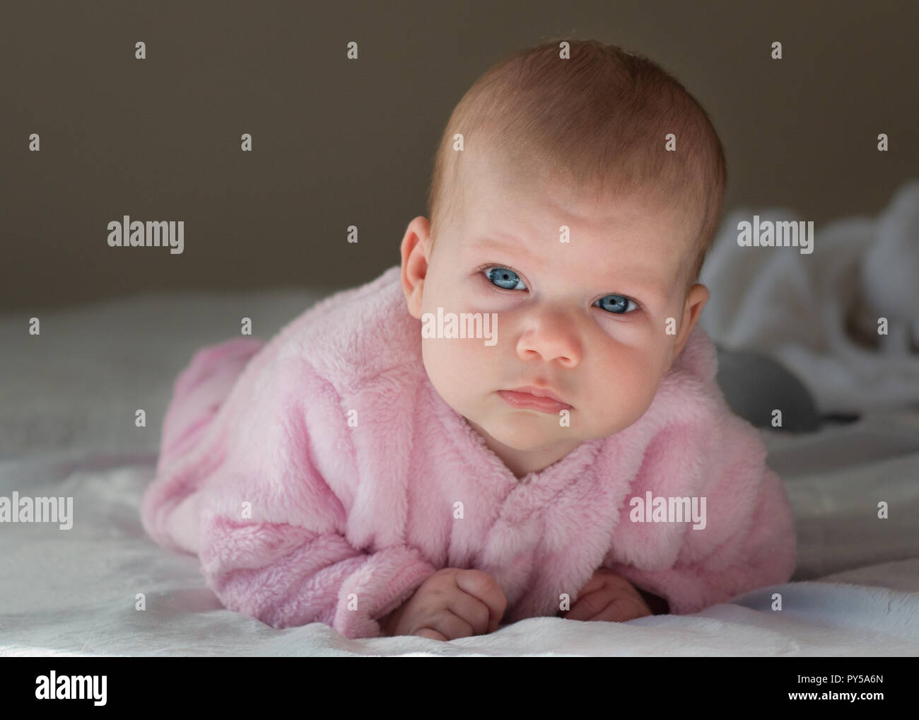 Baby girl laying on her stomach looking up at the camera with bright blue eyes wearing pink close up of her face. Stock Photo