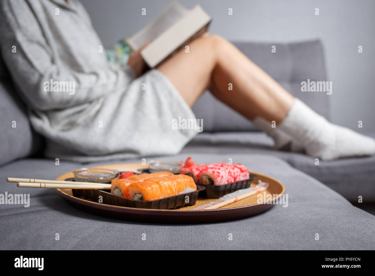 Young woman reading book and eating sushi on the sofa at home Stock Photo
