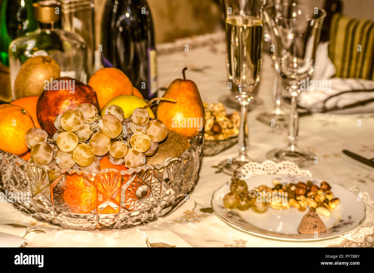 Crystal vase with fruit, glasses with champagne, different bottles, saucer with nuts, raisins and chocolate on a festive table Stock Photo