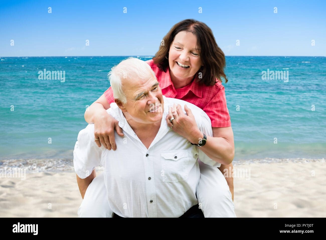 Portrait Of A Happy Senior Man Giving Piggyback To His Wife At Beach Stock Photo