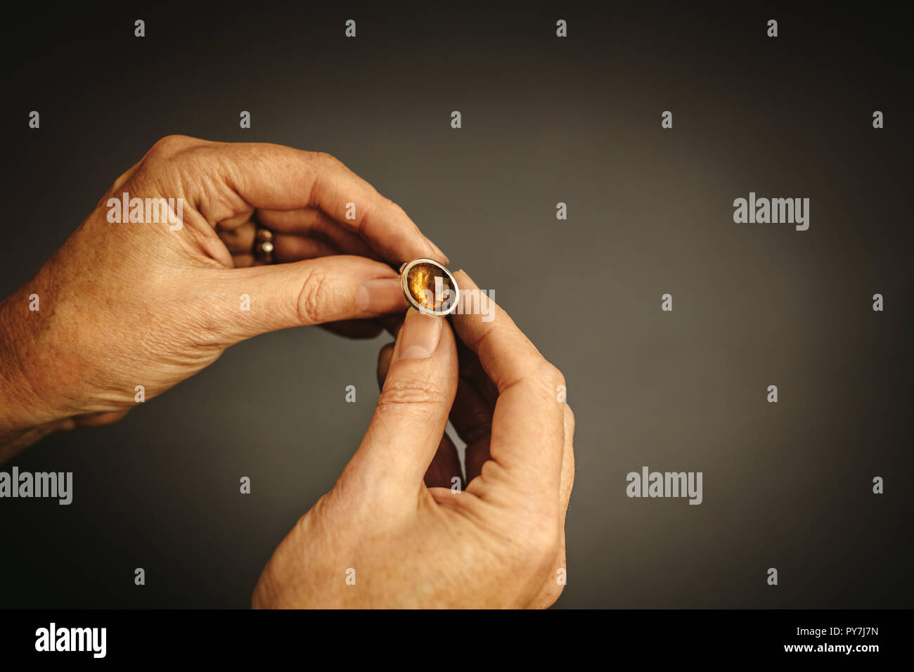Close up of female jewelry maker hands examining silver ring with gem stone against grey background. Jeweler inspecting ring. Stock Photo
